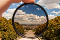Hand hält Filter, der Landschaft mit blauen Himmel und grünen Bäumen scharf erscheinen lässt.